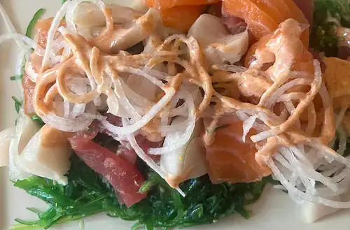 Seaweed salad with sashimi, spicy mayo, and vegetables on a white plate.