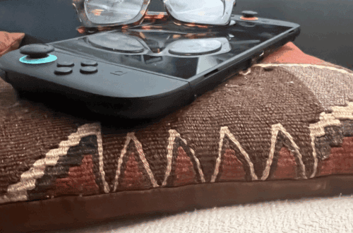 Close-up of a handheld gaming console with glasses resting on top, placed on a patterned pillow with earth tones and stitched detailing.