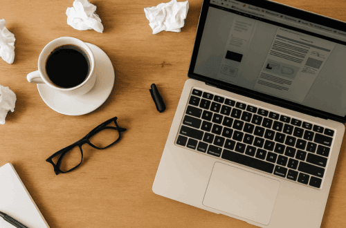 Top-down view of a laptop, black coffee, eyeglasses, pen, notebook, and crumpled paper on a wooden desk.