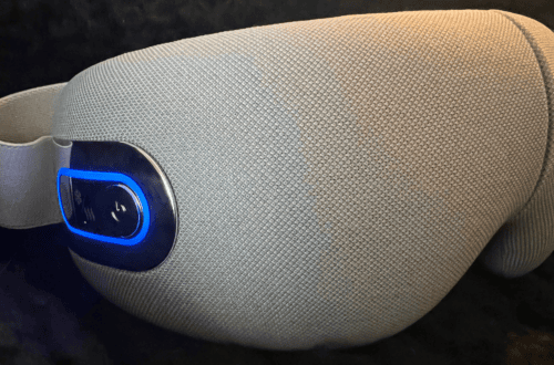 Close-up side view of a beige heated eye massager with a blue illuminated power button resting on a dark surface.