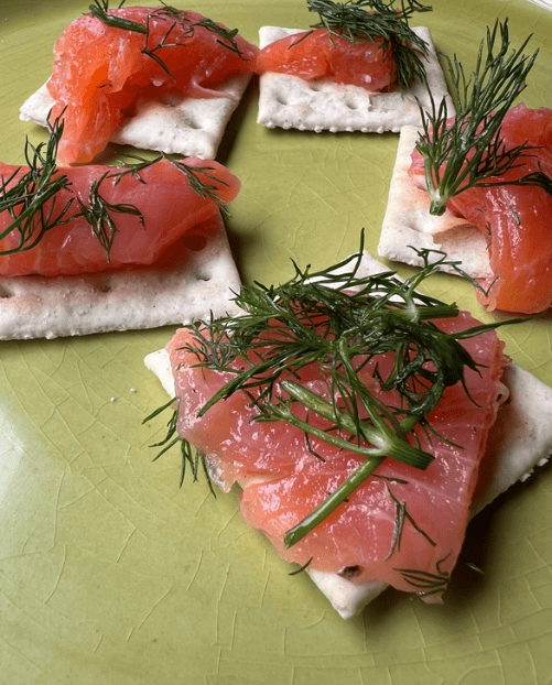 Square crackers topped with smoked salmon and fresh dill arranged on a green plate, creating a simple no-cook appetizer or light snack.