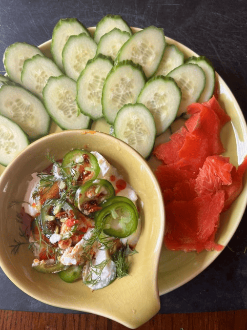 Plate with sliced cucumbers and smoked salmon served alongside a small bowl of creamy dip topped with fresh dill, sliced jalapeños, and red pepper flakes.