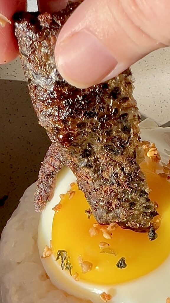 Close-up of grilled salmon skin atop a soft-boiled egg in a bowl.