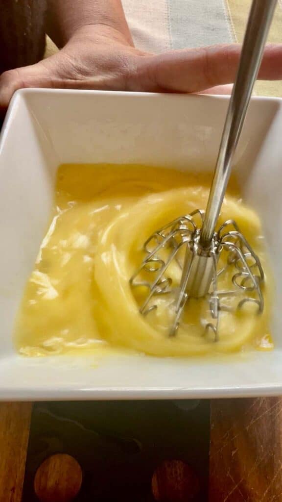 Close-up of a hand whisking a yellow creamy mixture in a white bowl.