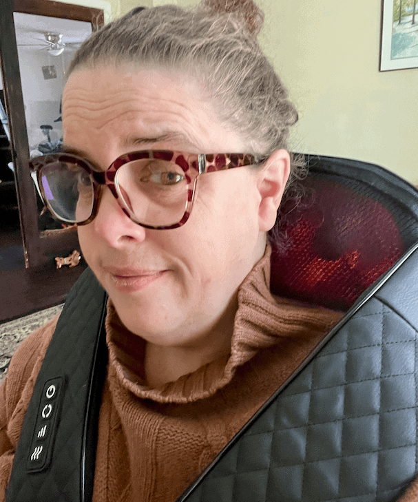 Woman with glasses and brown sweater sitting in a chair using a FSA eligible shoulder massager.