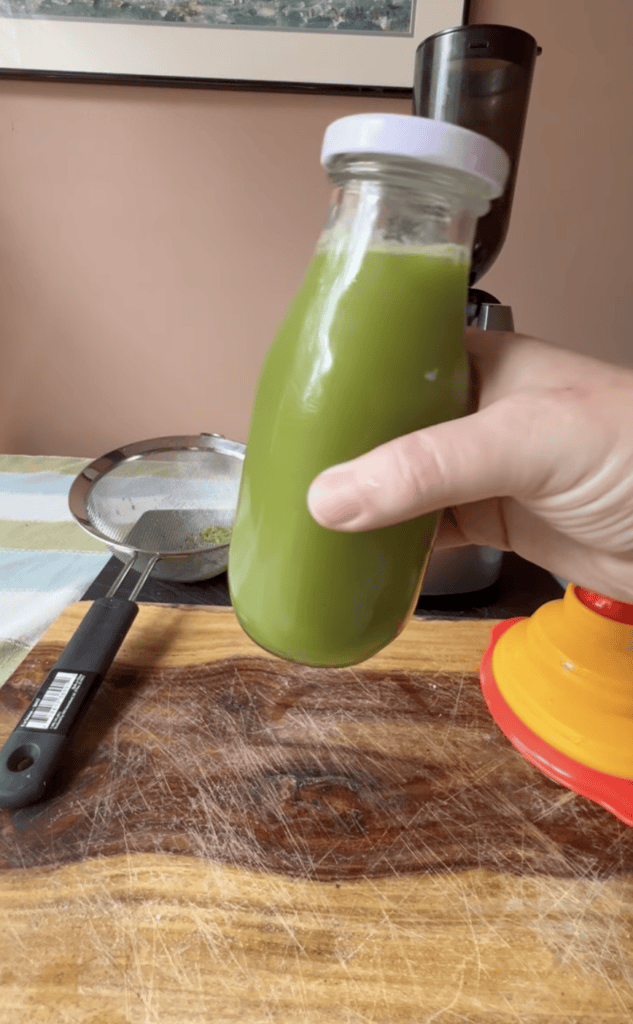 Hand holding a glass bottle filled with fresh green juice in a kitchen setting.
