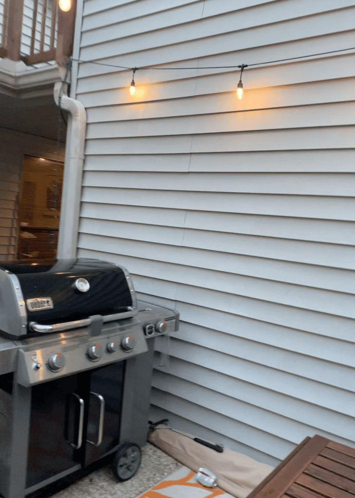 Outdoor patio with string lights and a barbecue grill against a house wall.