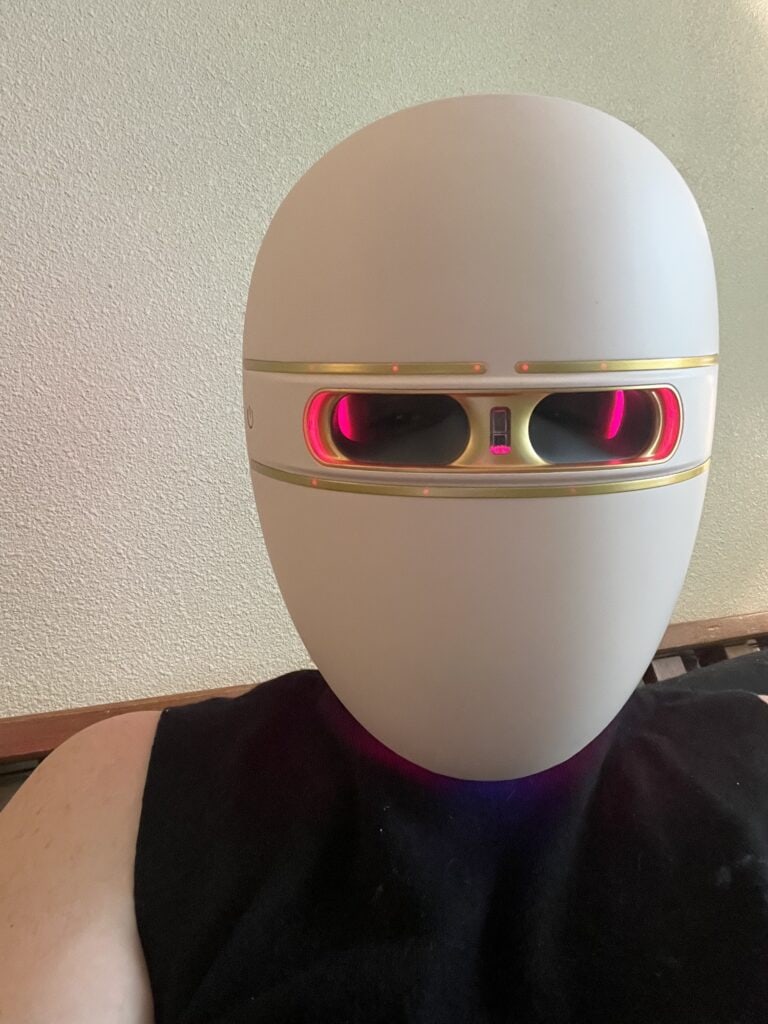 Close-up of a white facial light therapy mask with gold trim and glowing red LED lights around the eye openings, resting against a textured wall background.