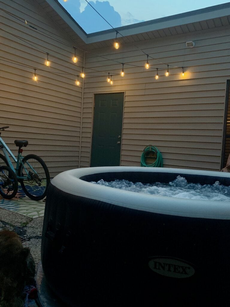 Outdoor hot tub under string lights with a green door and bicycle nearby.