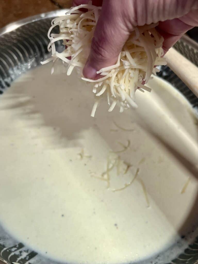 Hand sprinkling shredded cheese into a creamy soup in a metal bowl.