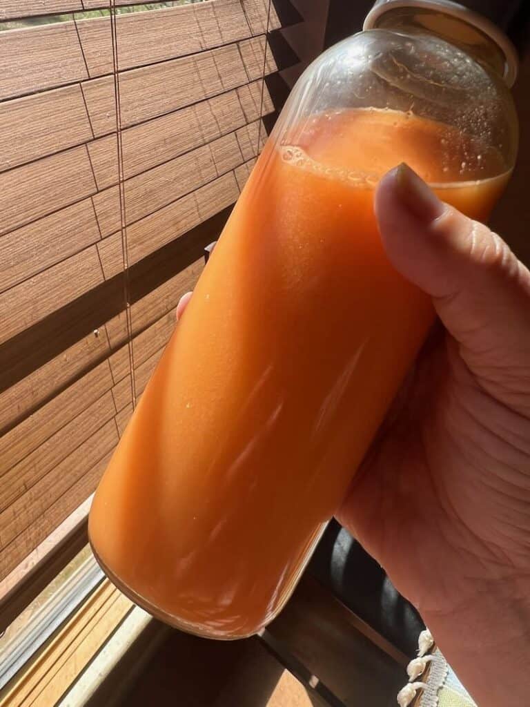 Fresh orange juice in a glass bottle held by hand near wooden blinds.