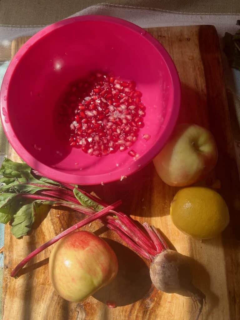 Fresh pomegranate seeds in a pink bowl with apples lemon and beet on cutting board.