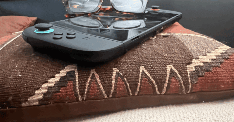 Close-up of a handheld gaming console with glasses resting on top, placed on a patterned pillow with earth tones and stitched detailing.