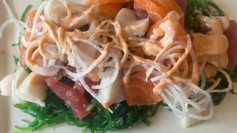 Seaweed salad with sashimi, spicy mayo, and vegetables on a white plate.