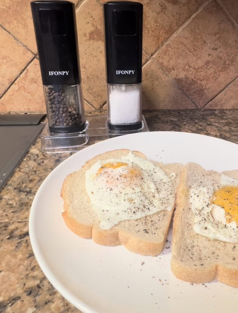 Two slices of bread with fried eggs on a white plate, salt and pepper shakers nearby.