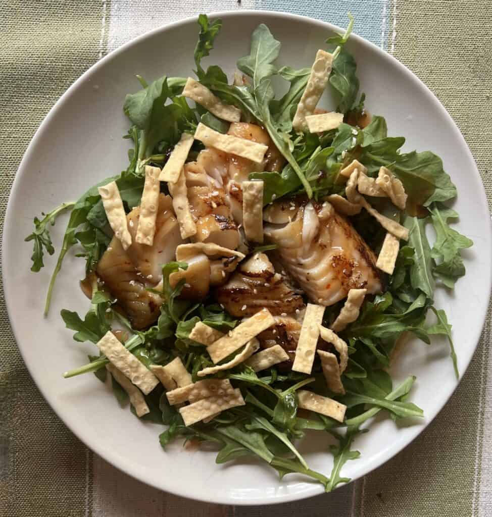 Healthy salad with miso glazed cod, arugula, and crispy breadsticks on a white plate.