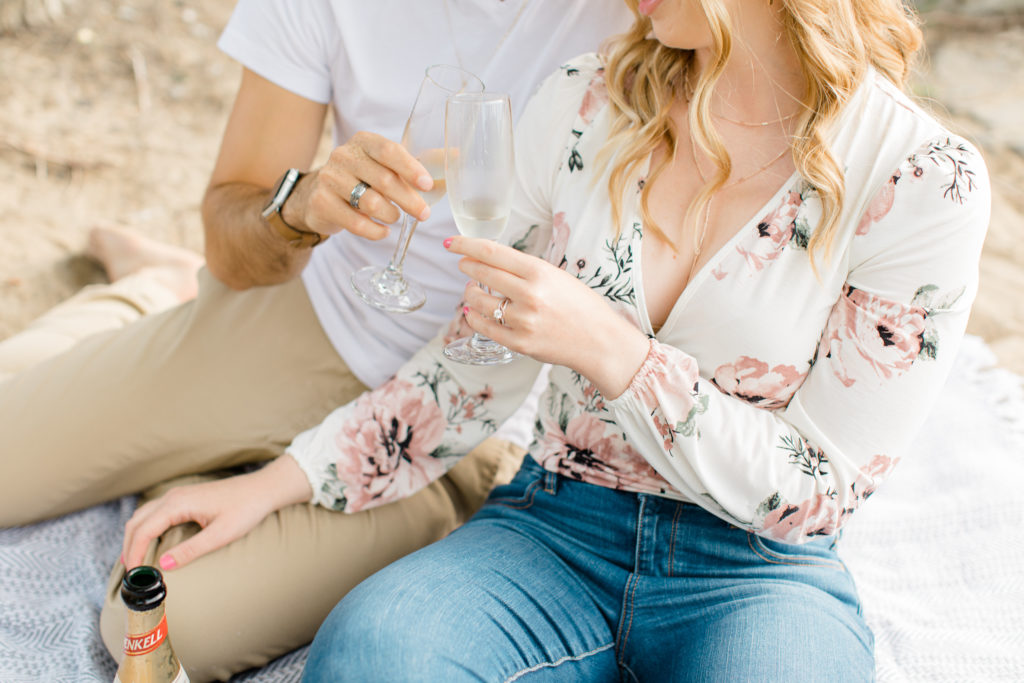 Champagne Cheers - Sitting on the Beach - Ideas for what to wear for Engagement Photography, Modern Engagement Session Inspiration Wardrobe Ideas. Unsure of what to wear for your engagement photos, we've got you! Romantic floral shirt & jeans.  White T-shirt & neutral pants . Boat Shoes and Fancy. beaded sandals. Engagement at Petrie Island, Orleans. Grey Loft Studio is Ottawa's Wedding and Engagement Photographer Videographer for Real couples, showcasing photos that are modern, bright, and fun.