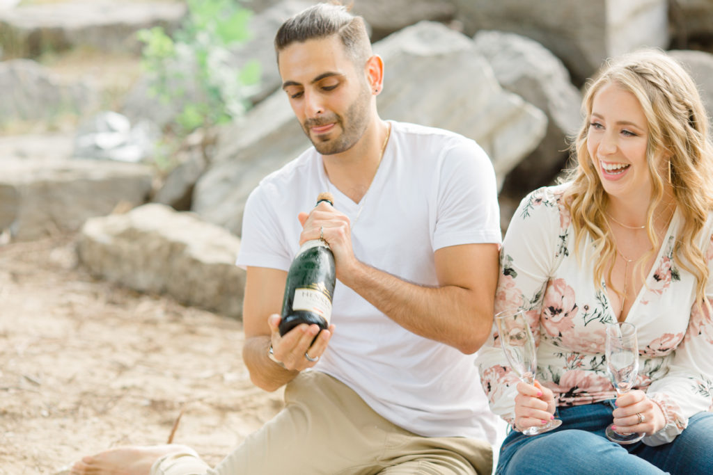 Shake the Champagne - Sitting on the Beach - Ideas for what to wear for Engagement Photography, Modern Engagement Session Inspiration Wardrobe Ideas. Unsure of what to wear for your engagement photos, we've got you! Romantic floral shirt & jeans.  White T-shirt & neutral pants . Boat Shoes and Fancy. beaded sandals. Engagement at Petrie Island, Orleans. Grey Loft Studio is Ottawa's Wedding and Engagement Photographer Videographer for Real couples, showcasing photos that are modern, bright, and fun.
