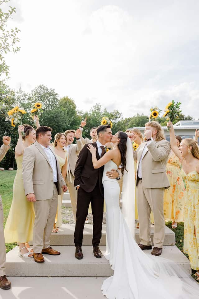 Julia and Will kissing on the steps as their wedding party cheers and raises sunflower bouquets