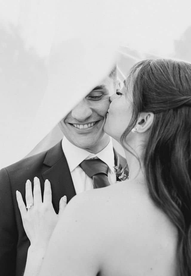 Black and white portrait of the bride kissing the groom's cheek under her veil