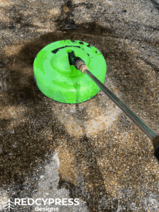A green surface cleaner pressure washing dirty concrete, revealing a bright clean path.