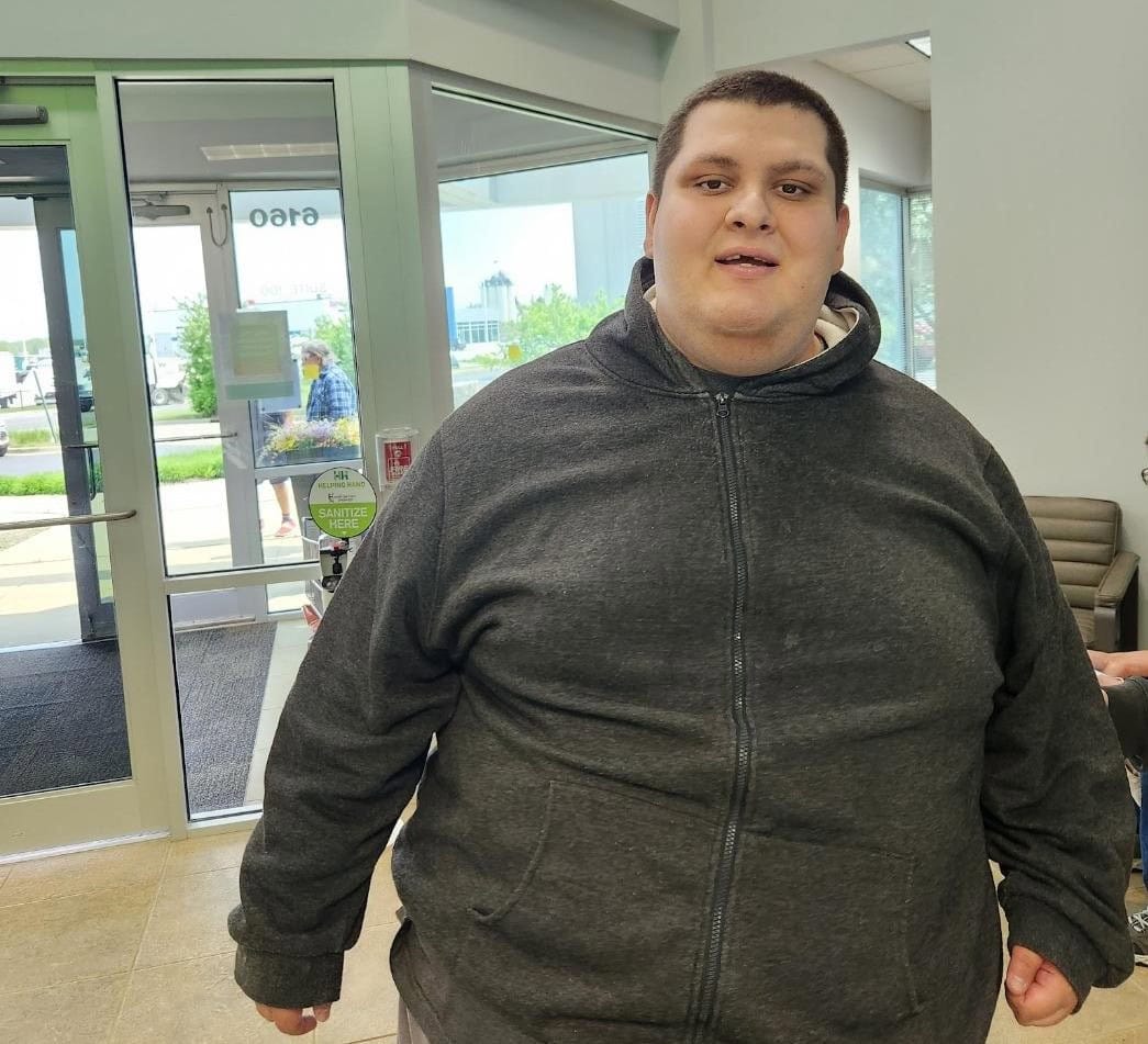 A person wearing a dark hoodie stands indoors near an exit door, smiling slightly. The area has glass doors leading outside and a few chairs are visible in the background. The scene appears to be in a public building or office.
