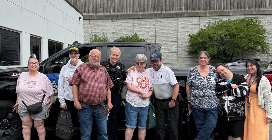 A group of nine people, including men and women of various ages, stand and smile in front of a black vehicle in a parking lot beside a white building under a partly cloudy sky.
