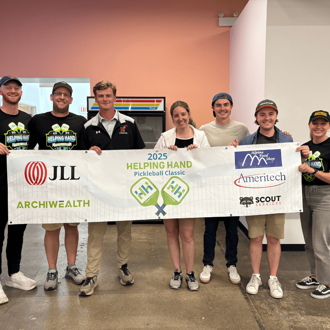 Seven smiling people stand indoors, holding a banner for the “2025 Helping Hand Pickleball Classic” with sponsor logos from JLL, ArchiWealth, Scout Services, and Ameritech. They are casually dressed and appear happy.