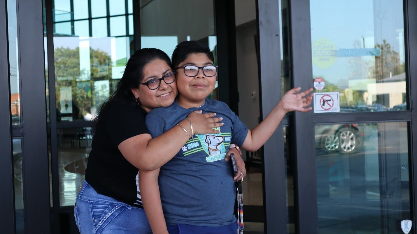 A woman hugs a young boy from behind as they stand outside a glass building entrance. The boy holds a lanyard and wears glasses and a graphic t-shirt. Both are smiling. Reflective glass reveals a bright day with blue sky and greenery.
