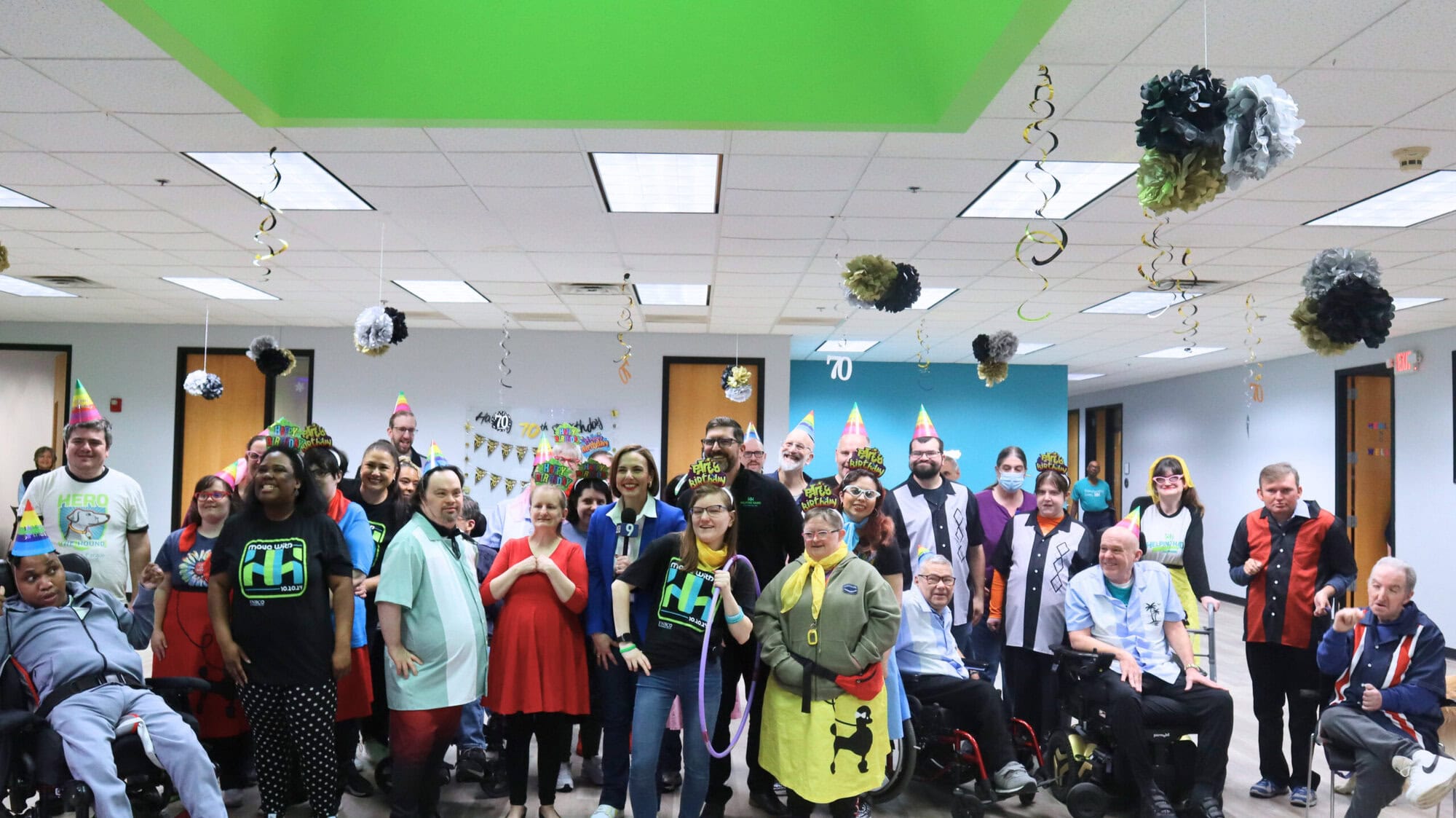 A diverse group of people, some in wheelchairs, pose together in a decorated room wearing party hats. Streamers and paper decorations hang from the ceiling, creating a festive atmosphere.