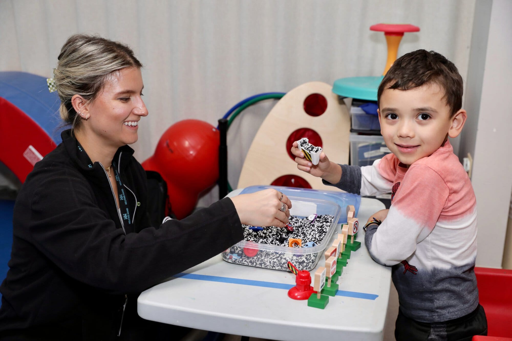 A woman and a young boy sit at a table playing with toys and sensory beads. The woman smiles at the boy, who looks toward the camera while holding a toy car. Colorful play equipment is visible in the background.