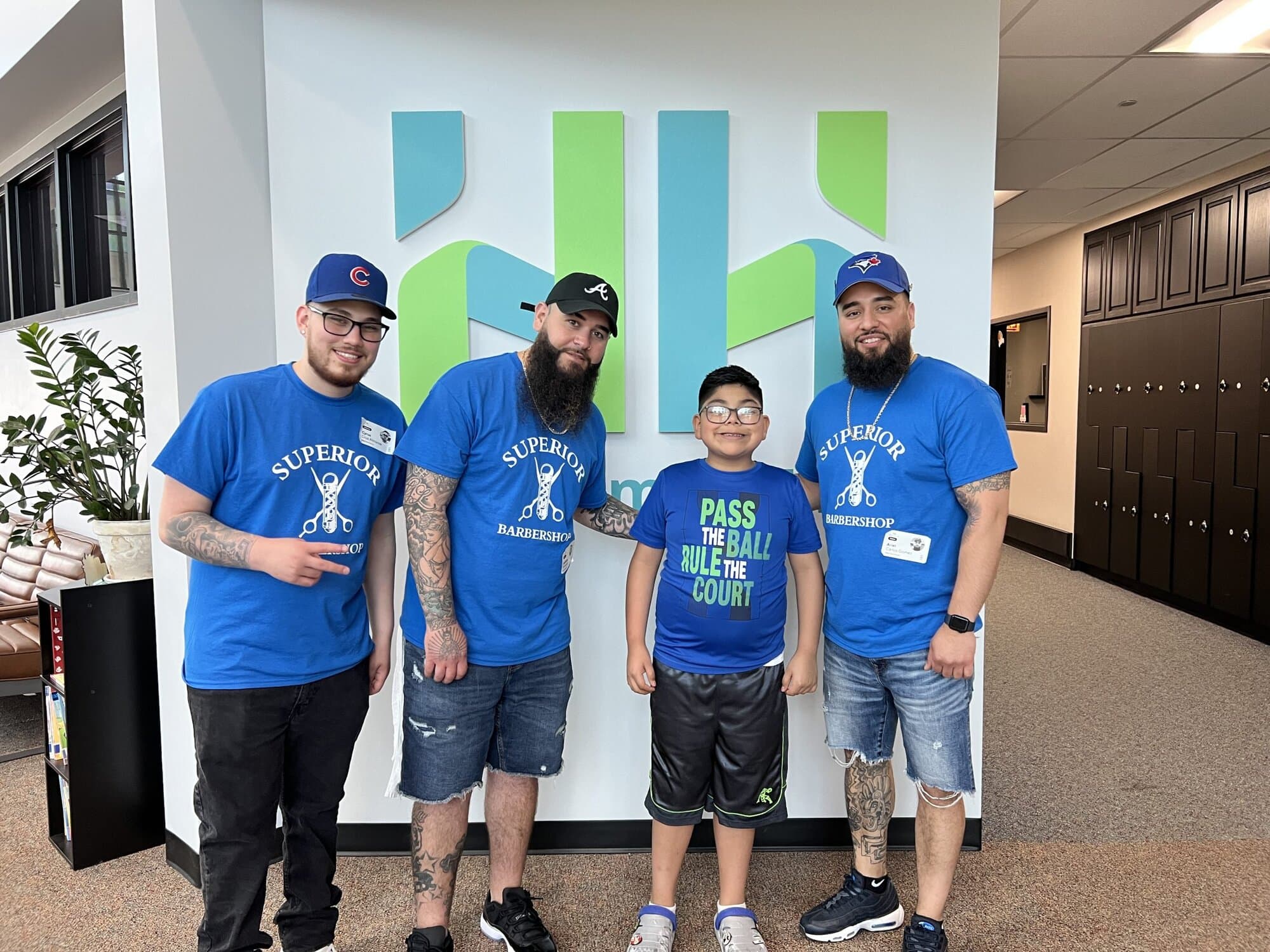 Three barbers in blue "Superior Barbershop" t-shirts stand with a smiling boy wearing a "Pass the Ball Rule the Court" shirt. They are in front of a green and blue abstract wall sign. The barbers are wearing hats and jeans, and the boy is in shorts and sneakers.