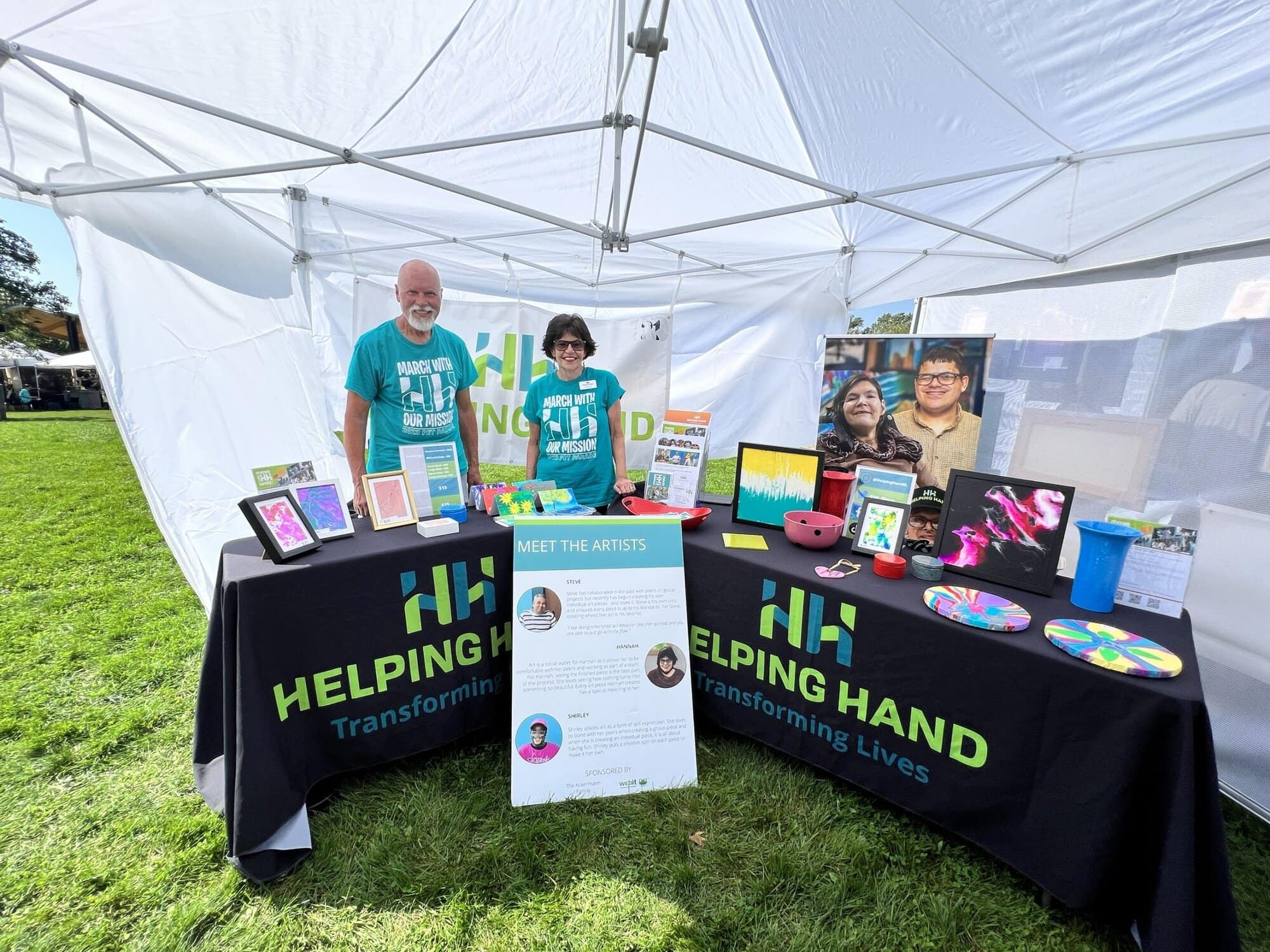Two people stand behind tables under a white canopy. The tables display artwork, photos, and flyers. Signs read "Helping Hand: Transforming Lives." The setting is outdoors on a grassy area.