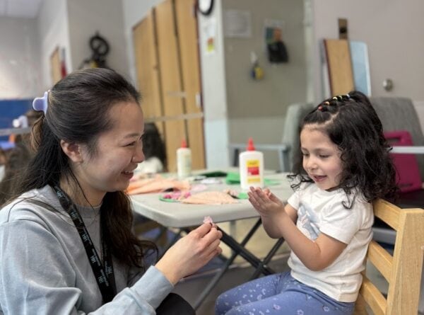 A woman kneels beside a smiling young girl sitting on a wooden chair. They are laughing together, with art supplies like glue and paper on the table behind them in a bright classroom setting.