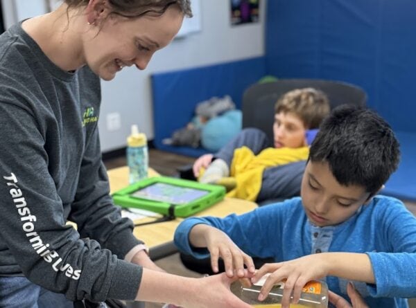 A smiling woman helps two children pour orange liquid from a measuring cup into a blender, while another child watches nearby in a classroom setting.