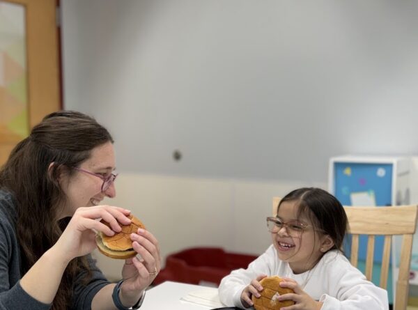 An adult and a young girl sit at a table, smiling at each other while holding burgers. Both have empty black plates in front of them. The setting appears to be a casual indoor space with neutral walls.
