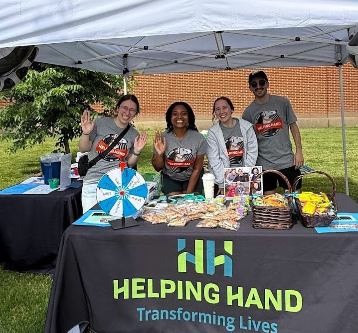 Four people stand and smile behind a booth table covered with snacks, flyers, and a prize wheel under a tent. The tablecloth reads “Helping Hand Transforming Lives.” Three people wave at the camera.