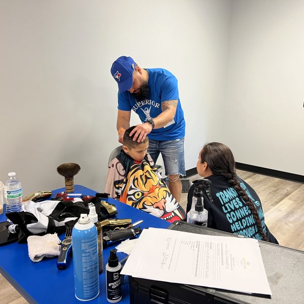 A barber in a blue cap and T-shirt gives a haircut to a young boy seated in a cape featuring a tiger design. A woman in a black jacket stands beside the boy, observing. The scene takes place in a bright room with a table of barber tools nearby.