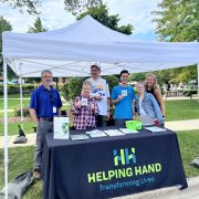 A group of five people stand under a white canopy tent, smiling and holding pamphlets. The table displays "Helping Hand: Transforming Lives" materials. The setting is outdoors with trees and grass in the background.