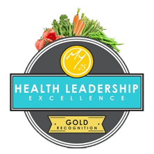 A circular badge with "Health Leadership Excellence" in the center, "Gold Recognition" below, and various vegetables like tomatoes, carrots, and greens at the top.