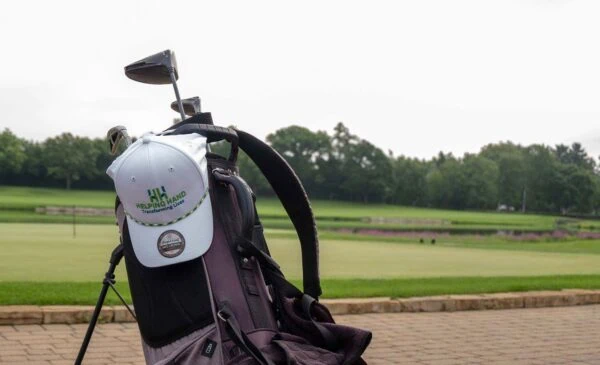 A golf bag with clubs and a white cap that says “HELPING HAND” is on a paved area next to a golf course, with green grass, trees, and a cloudy sky in the background.
