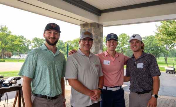 Four men stand together at a golf course, smiling for the camera. They wear golf attire, including collared shirts and hats. Trees, golf carts, and a grassy area are visible in the background.