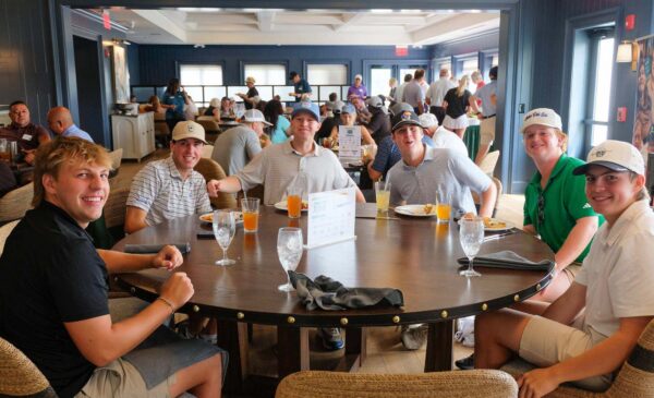 Six people sit around a round table in a restaurant, smiling at the camera. Drinks and menus are on the table. More diners and staff are visible in the background. The atmosphere is casual and bright.