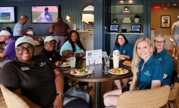 Six women sit around a restaurant table enjoying a meal, smiling at the camera. Four wear matching blue shirts and caps, while others are casually dressed. A TV and computer screens are visible in the background.