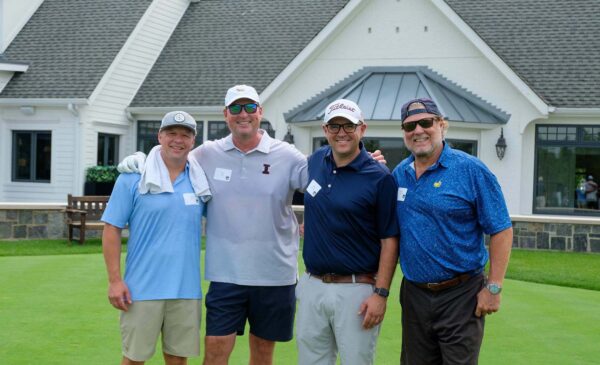 Four men in golf attire stand smiling on a putting green in front of a white clubhouse, arms around each other, wearing hats and sunglasses, with name tags on their shirts.