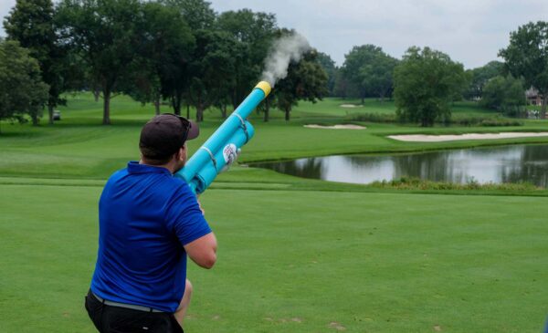 A person in a blue shirt and black cap fires a large blue air cannon on a golf course, with trees, sand traps, and a pond in the background. Smoke is visible from the cannon.