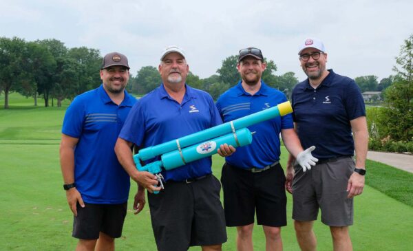 Four men in golf attire stand on a green golf course, smiling. One man holds a large blue and yellow toy-like launcher. Trees and a path are visible in the background.