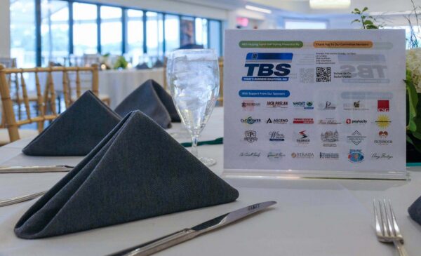 A formal table setting with folded napkins, a glass of water, and a display featuring business sponsor logos and event information in the background.