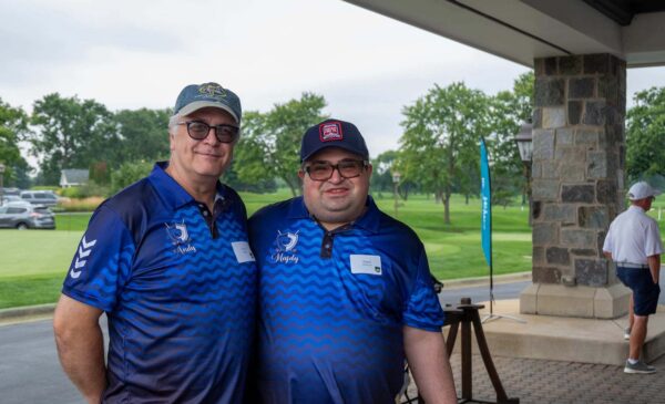 Two men wearing blue golf shirts and caps stand together and smile at an outdoor golf venue. A golfer is visible in the background near green grass and trees.