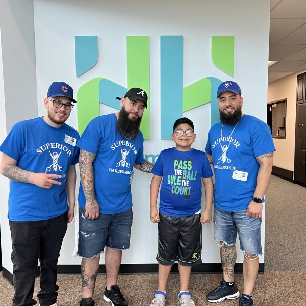Three barbers in blue "Superior Barbershop" t-shirts stand with a smiling boy wearing a "Pass the Ball Rule the Court" shirt. They are in front of a green and blue abstract wall sign. The barbers are wearing hats and jeans, and the boy is in shorts and sneakers.